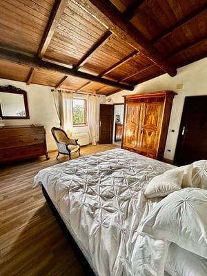 Spaziosa camera rustica con pavimento in legno chiaro, arredi classici e due finestre con tende bianche.