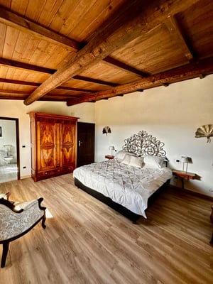 Camera matrimoniale luminosa con soffitto in legno a travi, letto con trapunta bianca, armadio e cassettone antichi.