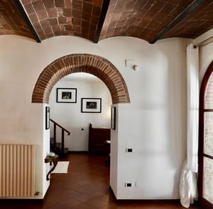 Corridoio con soffitto a volta in mattoni e arco in pietra che conduce a una stanza arredata con foto d’epoca.