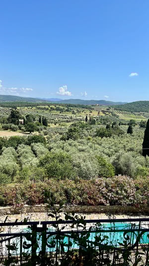 Veduta verticale su piscina e campagna toscana con cipressi e ulivi, sotto un cielo azzurro limpido.
